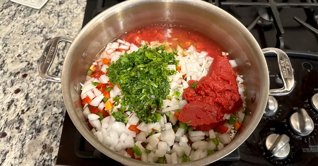 Tomato paste, tomatoes, onions, peppers, and cilantro in a stainless steel pot to make fresh vegetable salsa