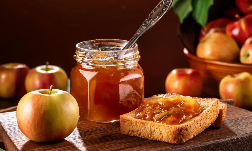 Canning jar of apple pie jam with a silver spoon sticking out of the top. Next to it on a cutting board are two pieces of toast with jam on them. There are apples in the background of the image.