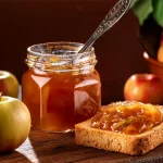 Canning jar of apple pie jam with a silver spoon sticking out of the top. Next to it on a cutting board are two pieces of toast with jam on them. There are apples in the background of the image.