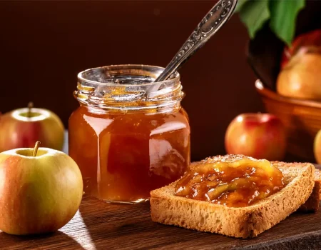 Canning jar of apple pie jam with a silver spoon sticking out of the top. Next to it on a cutting board are two pieces of toast with jam on them. There are apples in the background of the image.