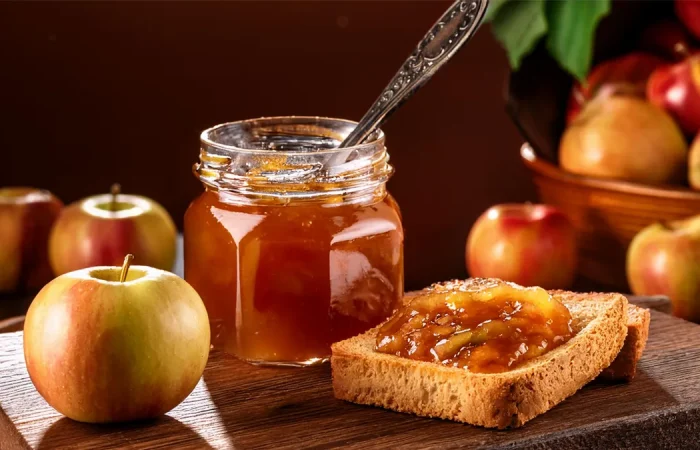 Canning jar of apple pie jam with a silver spoon sticking out of the top. Next to it on a cutting board are two pieces of toast with jam on them. There are apples in the background of the image.