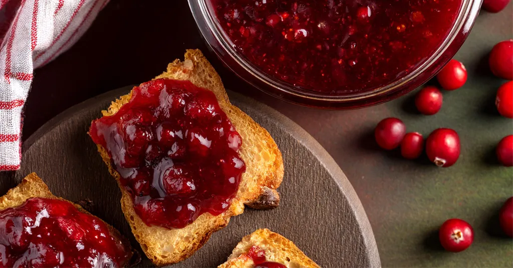 Three pieces of toast with cranberry raspberry jam spread on them along with a glass bowl of jam and scattered cranberries.