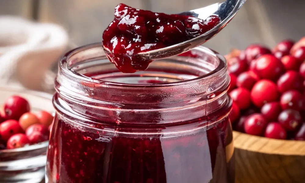 Mason jar of cranberry raspberry jam with a spoon full of jam. Two bowls of cranberries in the background.