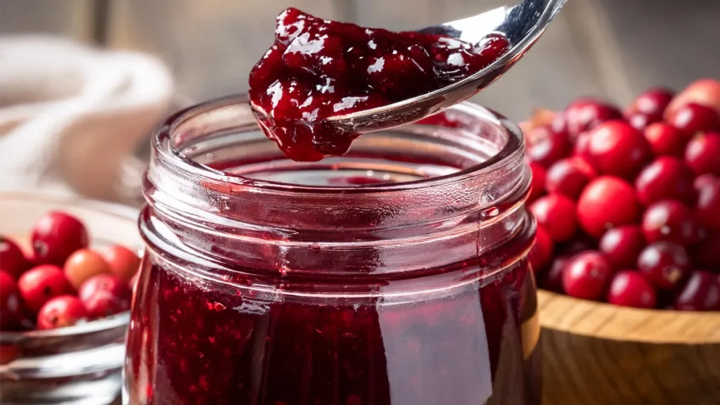 Mason jar of cranberry raspberry jam with a spoon full of jam. Two bowls of cranberries in the background.