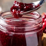 Mason jar of cranberry raspberry jam with a spoon full of jam. Two bowls of cranberries in the background.