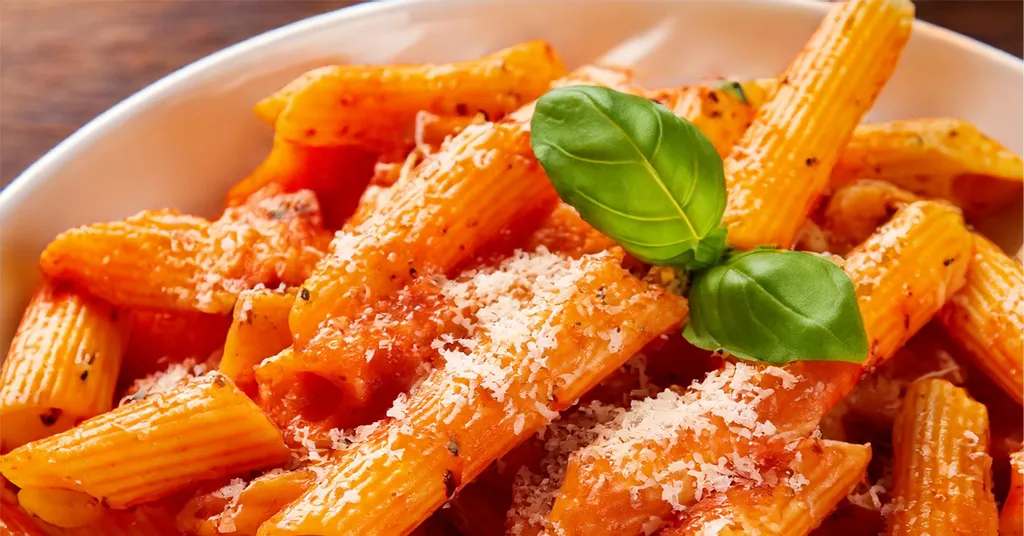 A close-up of penne pasta coated in creamy vodka sauce, topped with freshly grated Parmesan cheese and garnished with a basil leaf. The pasta is vibrant orange from the sauce, with visible seasoning and herbs.