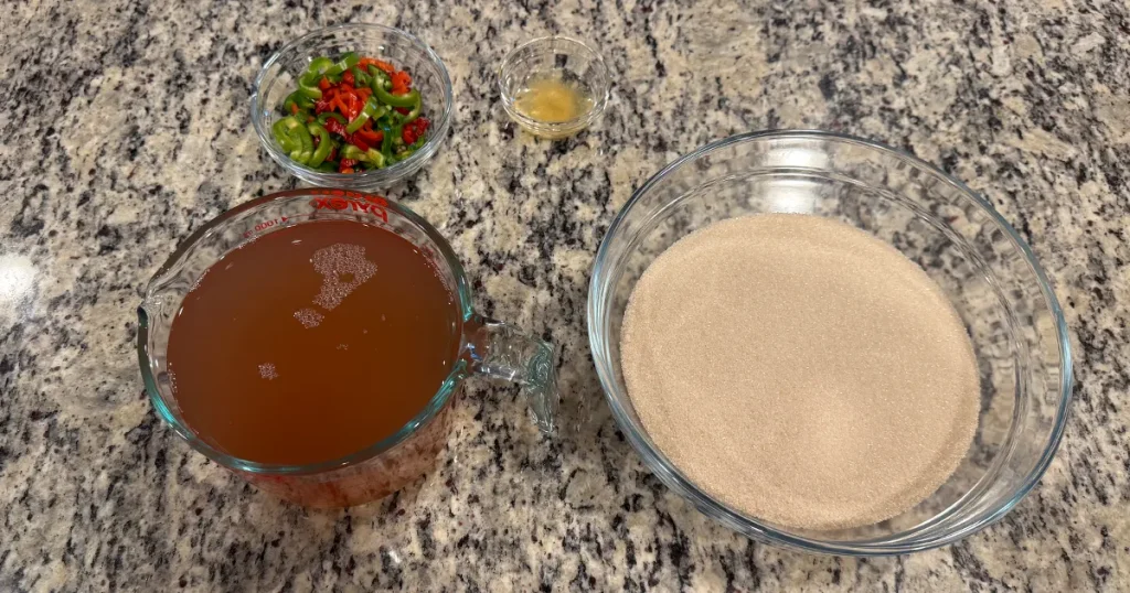 Glass bowls of apple juice, sugar, sliced jalapenos and lemon juice which are the ingredients for Apple Jalapeno Jelly.