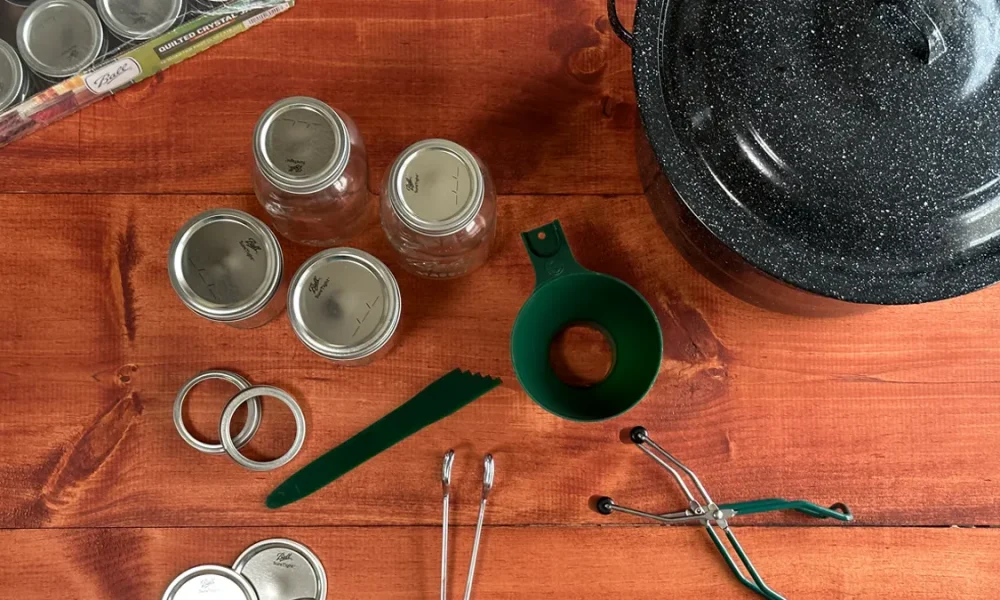 Canning Tools laid out on a wooden table. Canner, mason jars, funnel, headspace measurer, bands, lids, jar lifter, and tongs.