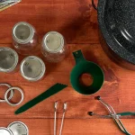 Canning Tools laid out on a wooden table. Canner, mason jars, funnel, headspace measurer, bands, lids, jar lifter, and tongs.