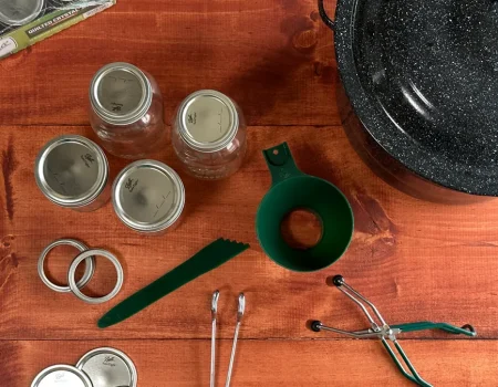 Canning Tools laid out on a wooden table. Canner, mason jars, funnel, headspace measurer, bands, lids, jar lifter, and tongs.