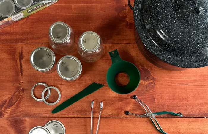 Canning Tools laid out on a wooden table. Canner, mason jars, funnel, headspace measurer, bands, lids, jar lifter, and tongs.