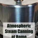 A close-up of a stainless steel atmospheric steam canner with a black knob on the lid, set on a stovetop. The text overlay reads 'Atmospheric Steam Canning at Home' with 'PreservingGuide.com,' making this image suitable for guides on steam canning techniques, home canning methods, and food preservation tutorials.