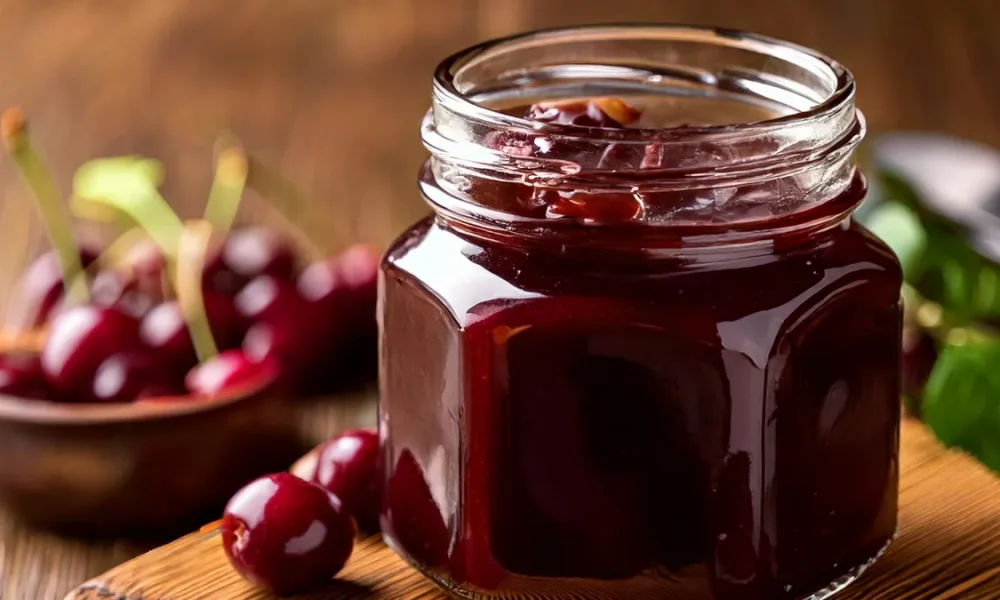 Chocolate cherry jam in a jar sitting on a wooden board with cherries in the background.