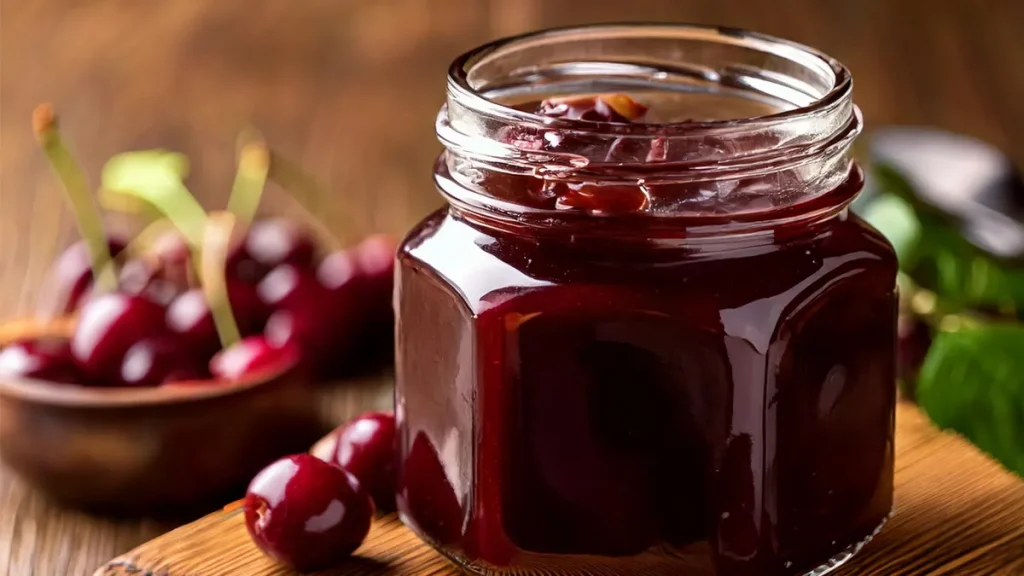 Chocolate cherry jam in a jar sitting on a wooden board with cherries in the background.