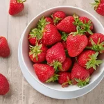 Ripe red strawberries in a white bowl with strawberries spread out on the counter around the bowl.