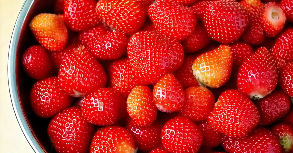 A close-up of a metal bowl filled with fresh, ripe strawberries. The strawberries are vibrant red with tiny seeds dotting their surface, and some have a slight gradient of orange near the top. The texture of the fruit's skin is glossy, reflecting light, and they appear freshly picked.