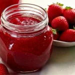 A glass jar filled with glossy, homemade strawberry jelly sits on a white surface. The deep red jelly has a slight texture. In the background, a white bowl is filled with fresh strawberries, and another jar of jam is partially visible.