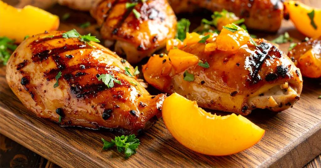 Grilled chicken breasts glazed with peach jelly are garnished with fresh herbs and diced peaches, served on a wooden board alongside peach slices.