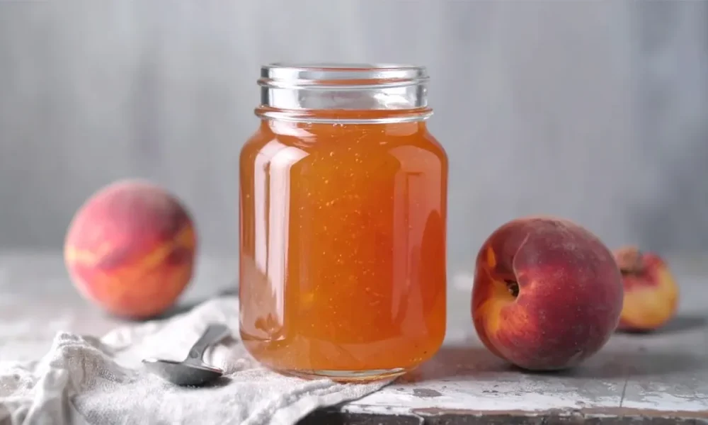 A glass jar filled with golden peach jelly sits on a rustic surface, flanked by two fresh peaches. A spoon rests on a light cloth beside the jar, adding a homemade, cozy touch.