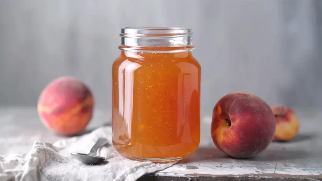 A glass jar filled with golden peach jelly sits on a rustic surface, flanked by two fresh peaches. A spoon rests on a light cloth beside the jar, adding a homemade, cozy touch.