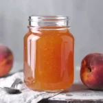 A glass jar filled with golden peach jelly sits on a rustic surface, flanked by two fresh peaches. A spoon rests on a light cloth beside the jar, adding a homemade, cozy touch.