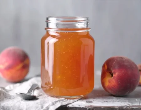A glass jar filled with golden peach jelly sits on a rustic surface, flanked by two fresh peaches. A spoon rests on a light cloth beside the jar, adding a homemade, cozy touch.