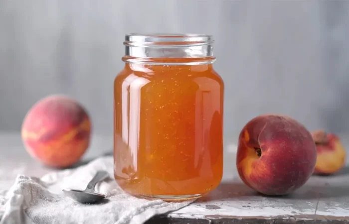 A glass jar filled with golden peach jelly sits on a rustic surface, flanked by two fresh peaches. A spoon rests on a light cloth beside the jar, adding a homemade, cozy touch.