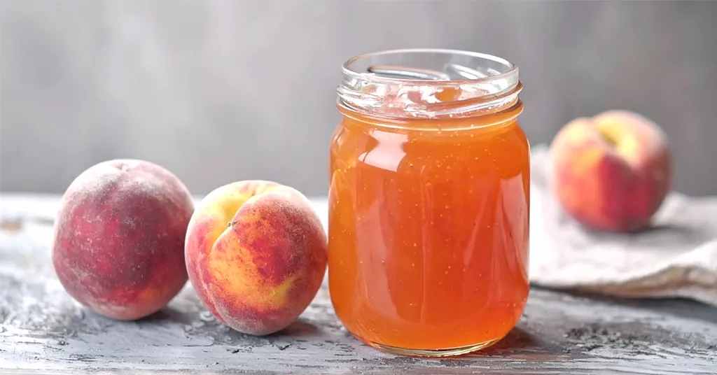 A clear glass jar filled with golden-orange peach jelly sits on a weathered surface, surrounded by three fresh peaches, with a soft cloth in the background for a rustic feel.