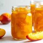 Glass jars filled with thick, golden peach pie filling are surrounded by fresh peaches and peach slices on a white countertop, showcasing a homemade canning recipe.