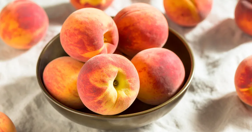 A rustic bowl filled with ripe, fuzzy peaches sits on a white linen cloth, surrounded by more peaches, ideal fruit for making a homemade peach pie filling recipe.