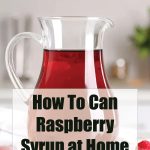 A clear glass pitcher filled with vibrant red raspberry syrup sits on a white kitchen counter, surrounded by a few fresh raspberries. Text overlay reads “How To Can Raspberry Syrup at Home – PreservingGuide.com.”