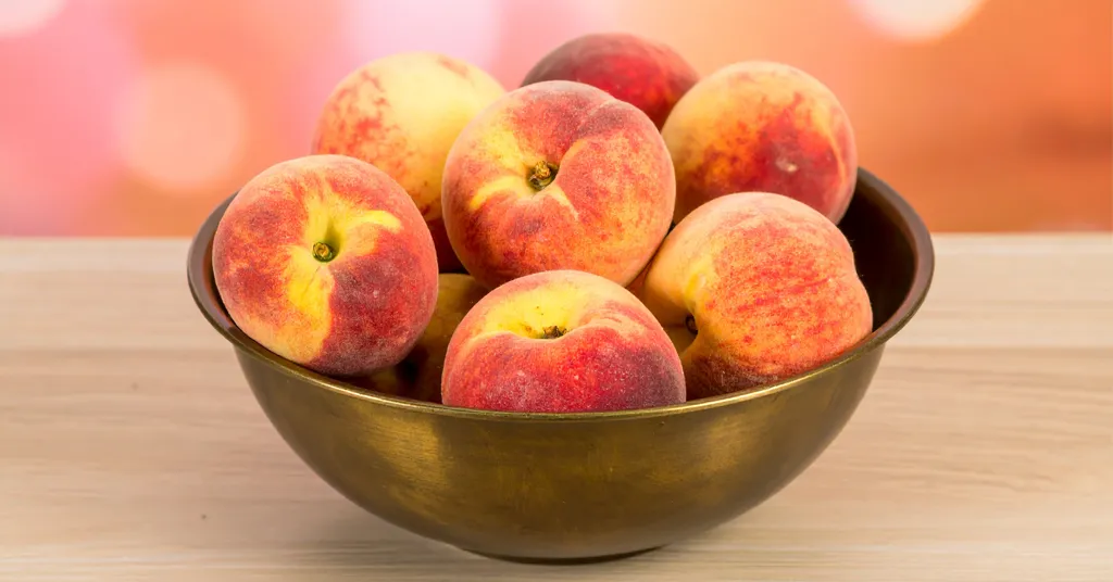Fresh picked peaches in a gold bowl sitting on a wooden surface.