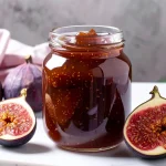 A glass jar filled with thick, dark fig preserves sits on a marble surface beside fresh whole figs and halved figs showing their rich red interiors. Perfect visual for a homemade fig preserves recipe highlighting ripe fruit and a rustic presentation.