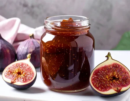 A glass jar filled with thick, dark fig preserves sits on a marble surface beside fresh whole figs and halved figs showing their rich red interiors. Perfect visual for a homemade fig preserves recipe highlighting ripe fruit and a rustic presentation.