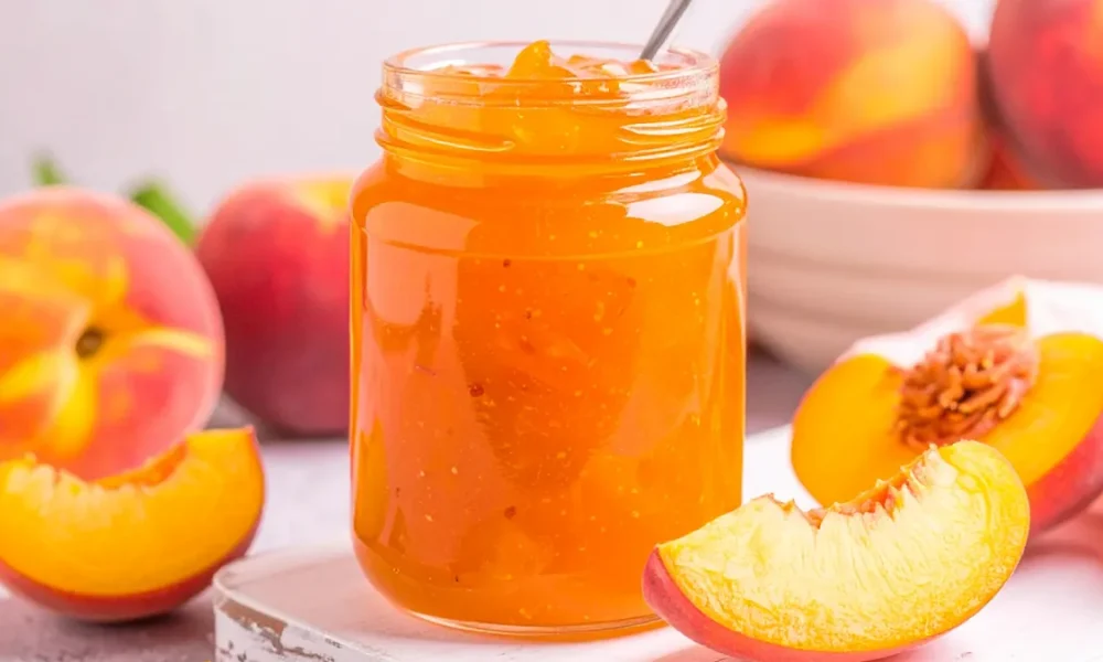 Peach Jam in a mason jar with sliced peaches and a bowl of peaches in the background.