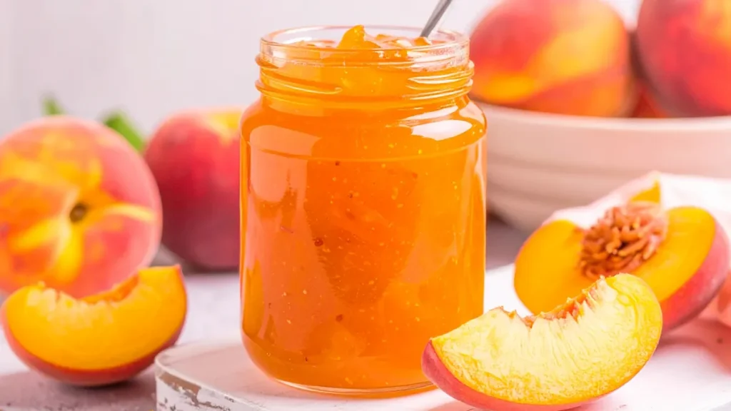 Peach Jam in a mason jar with sliced peaches and a bowl of peaches in the background.