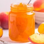 Peach Jam in a mason jar with sliced peaches and a bowl of peaches in the background.