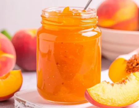 Peach Jam in a mason jar with sliced peaches and a bowl of peaches in the background.