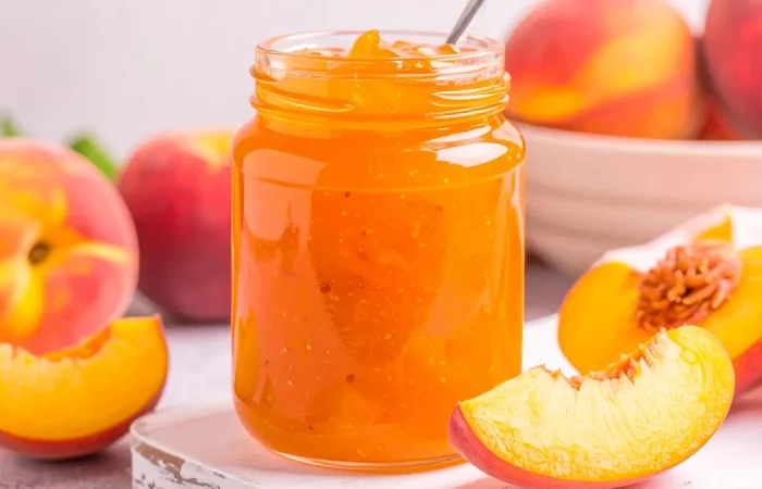 Peach Jam in a mason jar with sliced peaches and a bowl of peaches in the background.