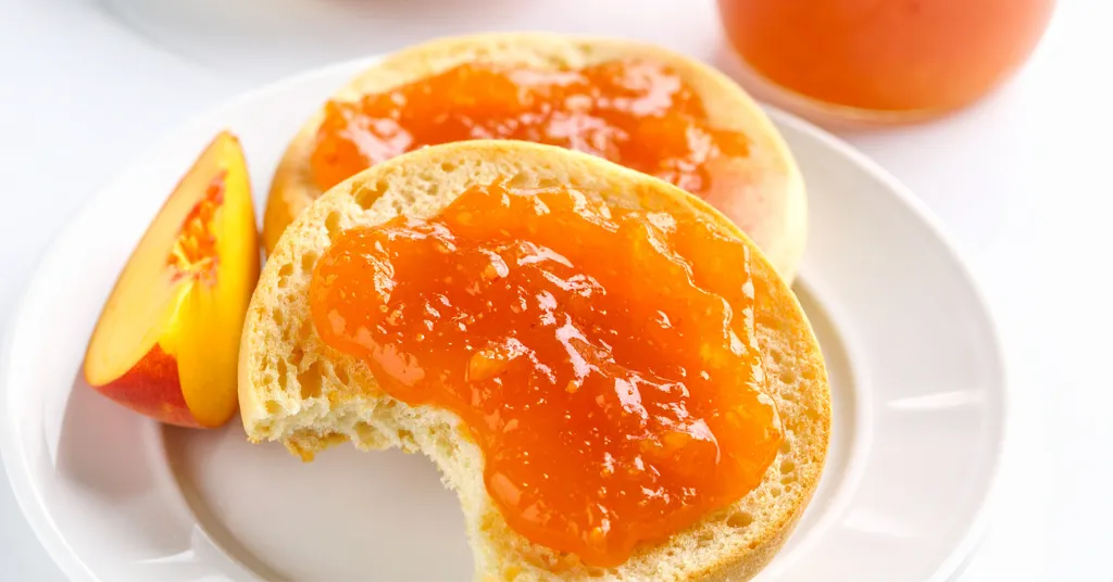 Peach jam spread on round toast on a white plate with a slice of peach.
