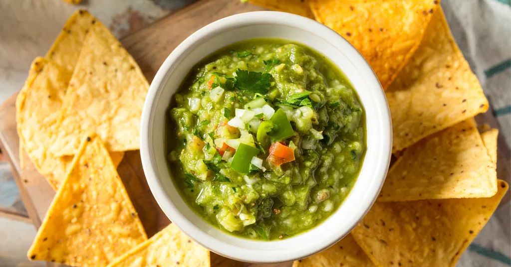Chunky Green Tomatillo Salsa with onions and jalapenos in a white bowl in the middle of tortilla chips on a cutting board.